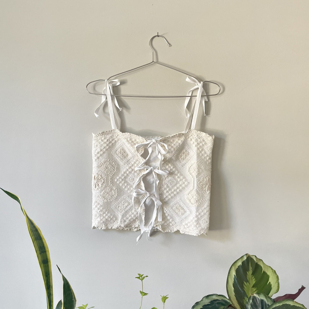 A Cream Crochet Festival Cropped Tank Top w/ Tie Back hanging on a wall next to a potted plant.