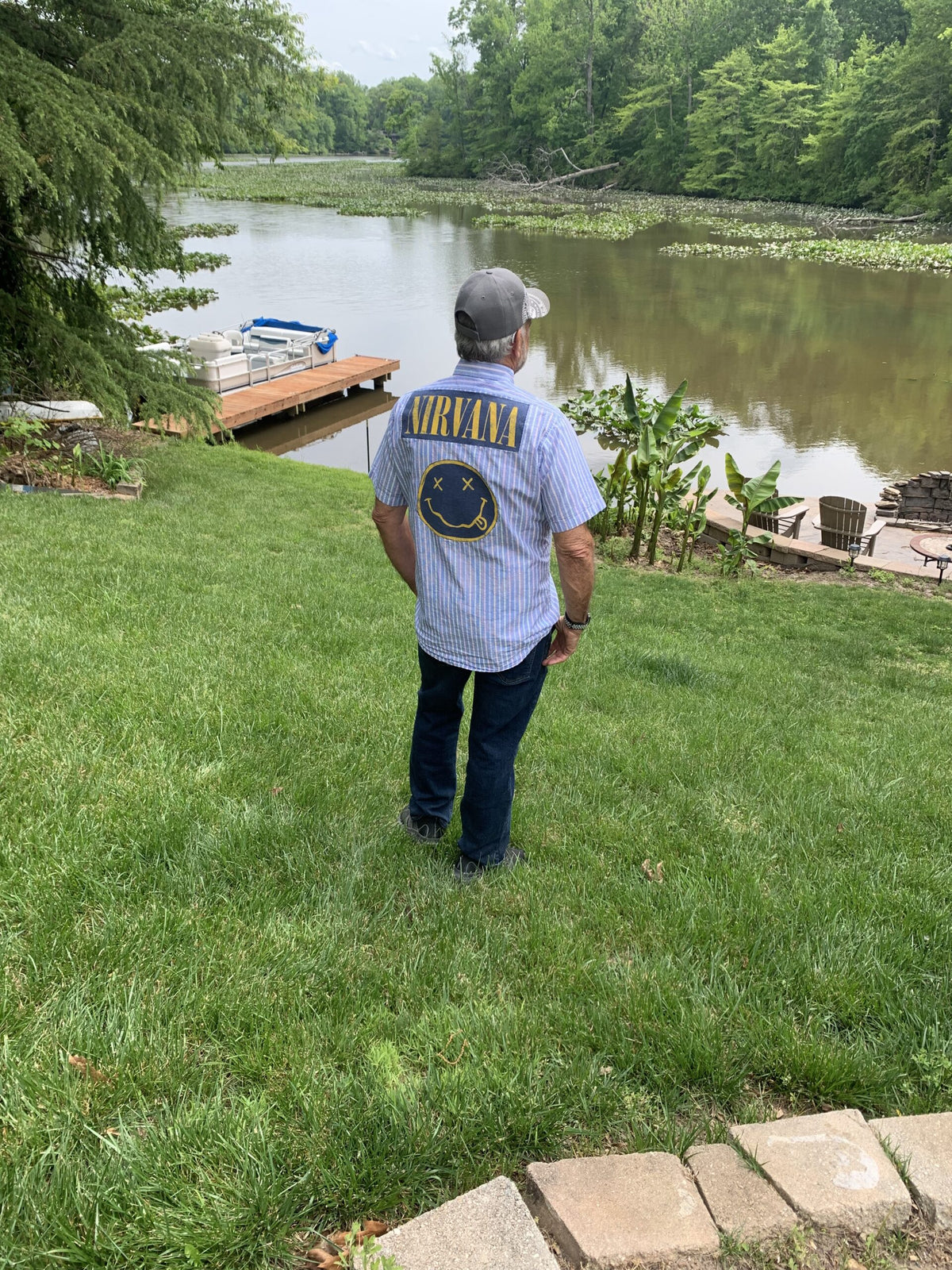 a man wearing Nirvana mens pinstriped button up shirt standing on top of a lush green field next to a lake.