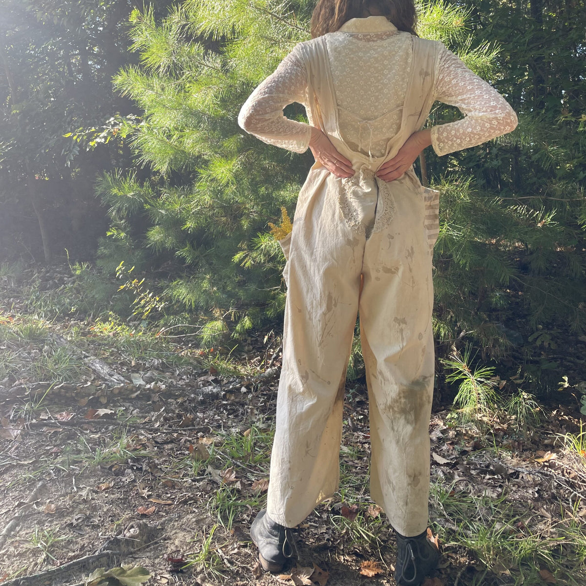 A woman is standing in the woods wearing overalls.