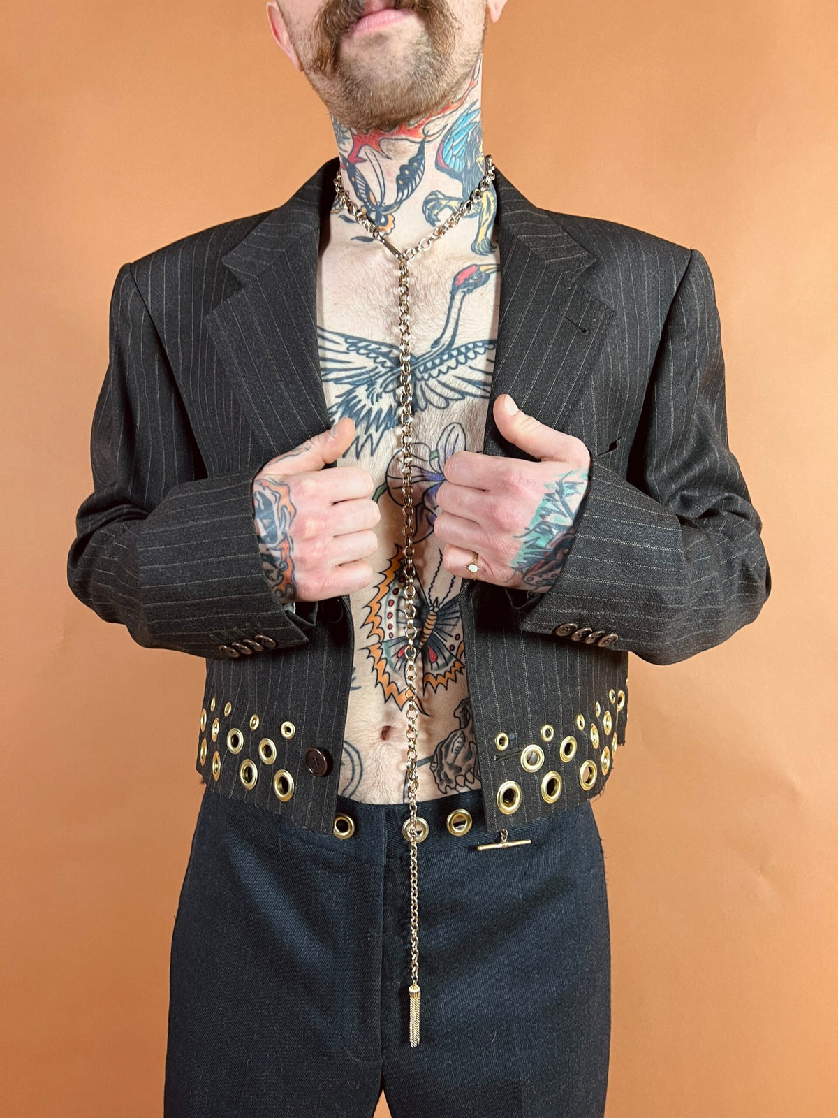 A bearded man in an open pinstripe jacket with studded lapels reveals an array of chest tattoos against a tan background.