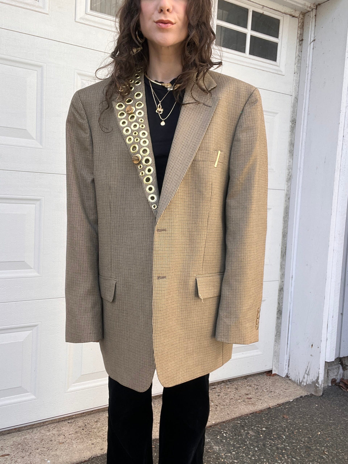 A person wearing an oversized houndstooth blazer and black trousers stands in front of a white garage door.