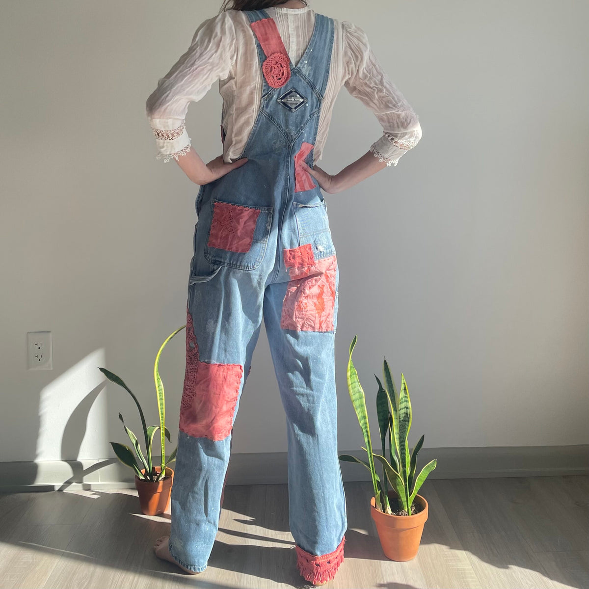 A woman is standing in front of a potted plant wearing denim overalls.