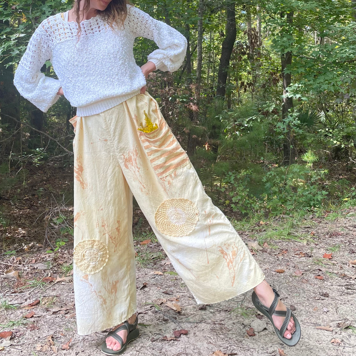 A woman is standing in the woods wearing yellow wide leg pants.