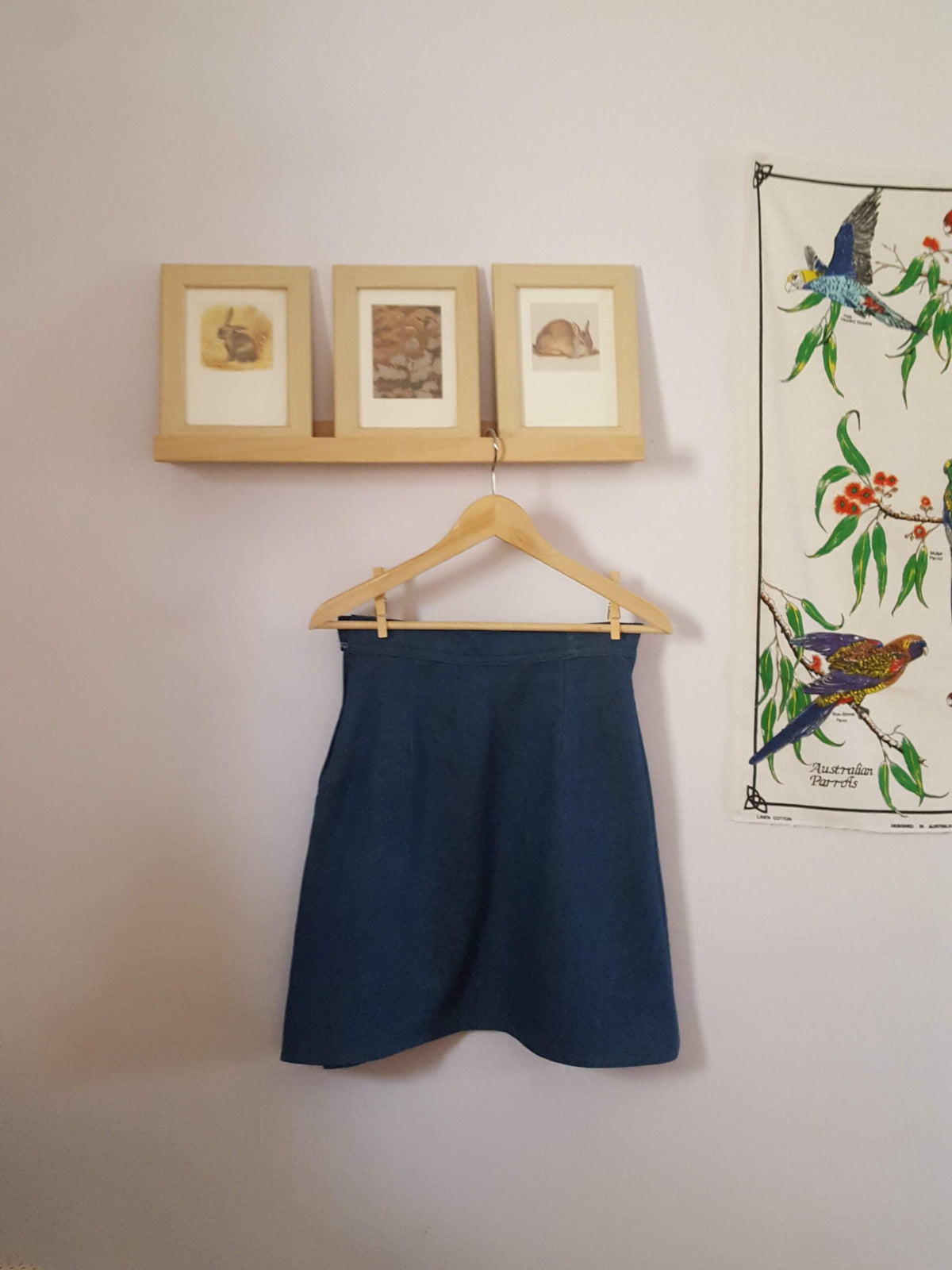 A pair of framed pictures hang on a wall next to an Embroidered Dark Wash Kangaroo Pocket Mini Skirt hanging on a hook.