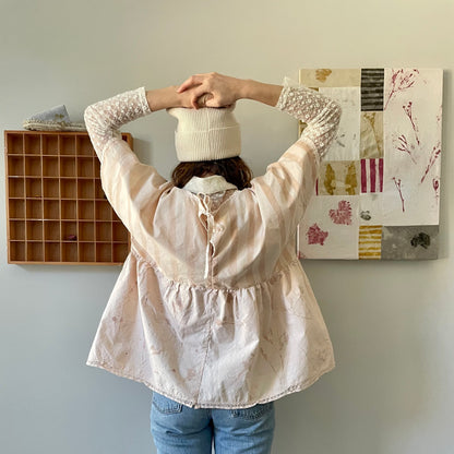A woman is standing in front of a wall with a hat on her head. She is wearing an avocado naturally dyed pink top.