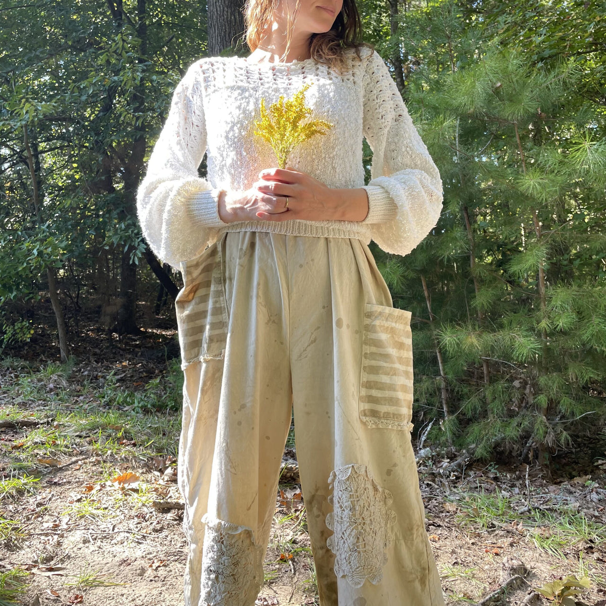 A woman is standing in the woods wearing a sweater and pants.