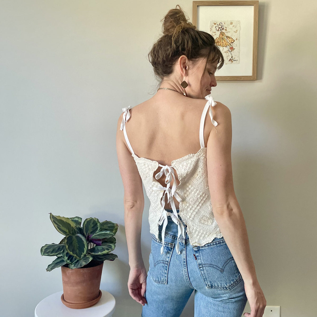 A woman in a Cream Crochet Festival Cropped Tank Top w/ Tie Back is standing next to a potted plant.