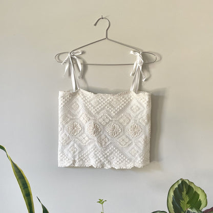 A Cream Crochet Festival Cropped Tank Top w/ Tie Back hanging on a wall next to a potted plant.