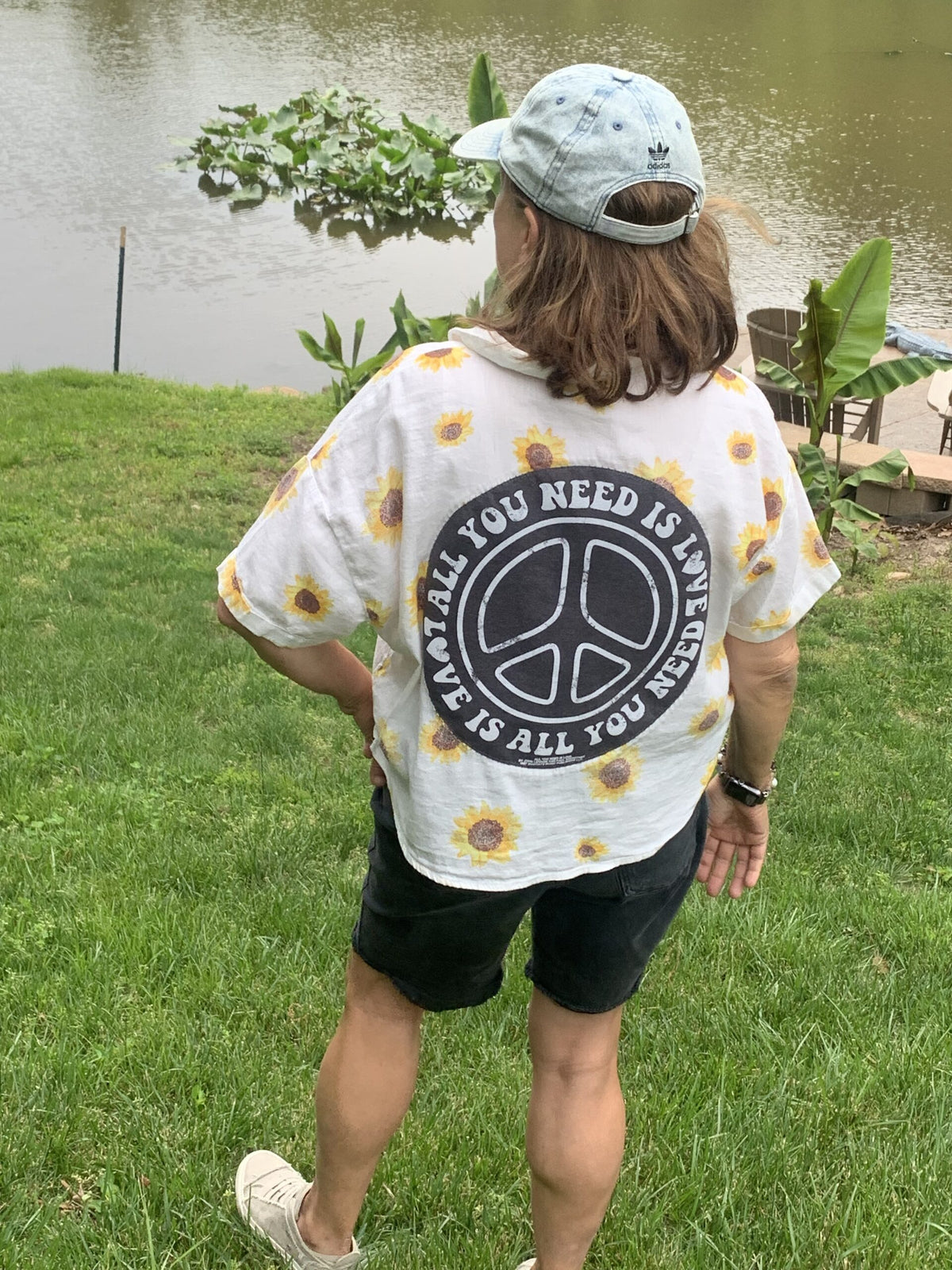 A person standing in the grass near a body of water wearing an All You Need Is Love - Sunflower button up shirt.