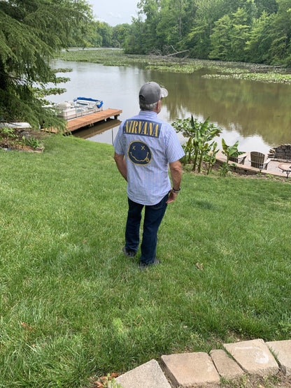 a man wearing Nirvana mens pinstriped button up shirt standing on top of a lush green field next to a lake.
