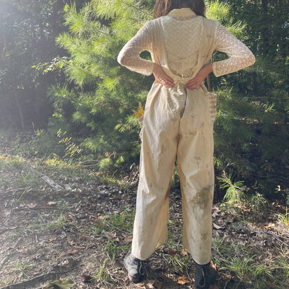 A woman is standing in the woods wearing overalls.