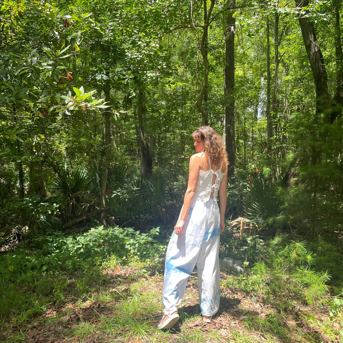 A woman standing in the woods wearing a white jumpsuit.