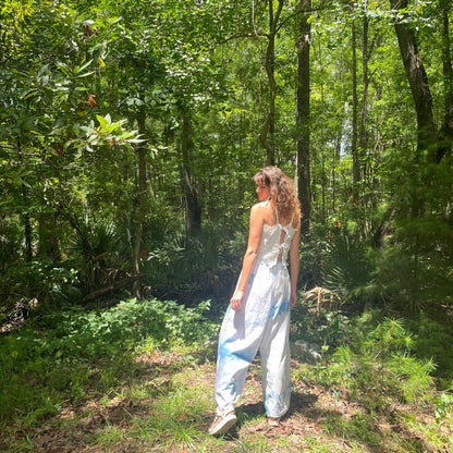A woman standing in the woods wearing a white jumpsuit.