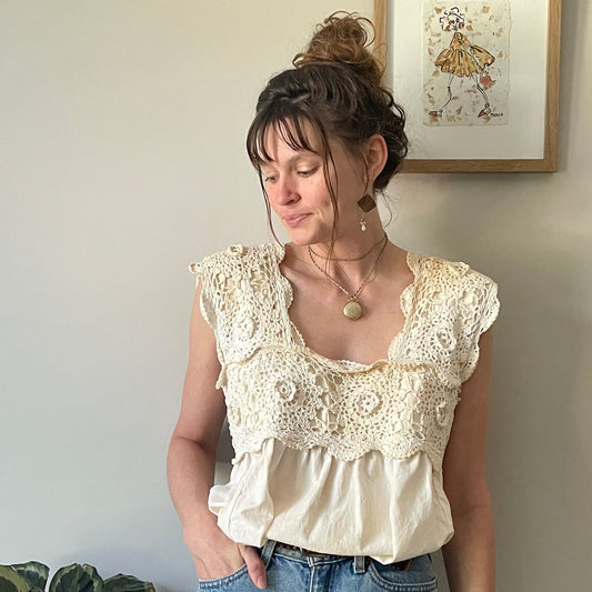 A woman standing in front of a white wall wearing the Tea Stained Cream Crochet Festival Tank Top w/ Tie Back.