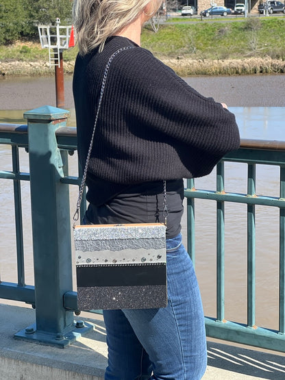Woman leaning on a railing overlooking a river, wearing a black sweater and jeans, with a silver-striped shoulder bag.