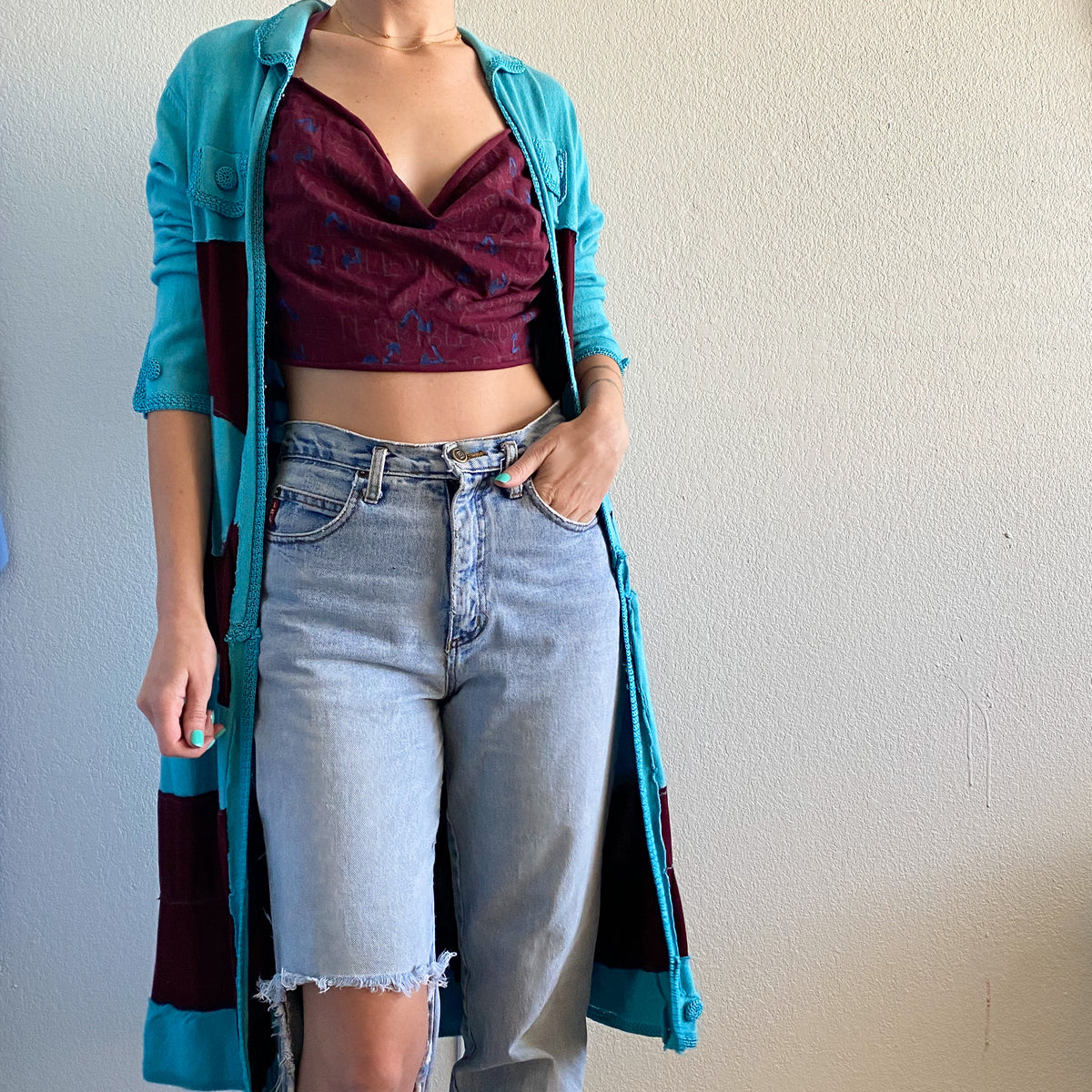 A woman standing in front of a white wall wearing a 1of1 painted text print maroon halter crop top.