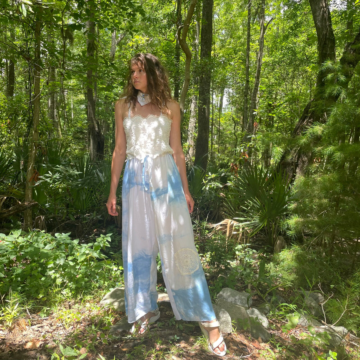 A woman is standing in the woods wearing a tie dye jumpsuit.
