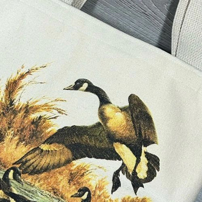 Image of a tote bag featuring a detailed illustration of a canada goose in a dynamic pose among tall grasses.