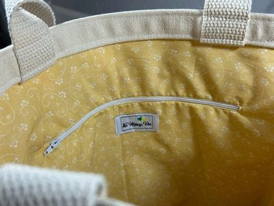 Close-up view of a yellow fabric tote bag with a zippered pocket and decorative stitching, featuring a branded label sewn on the interior.