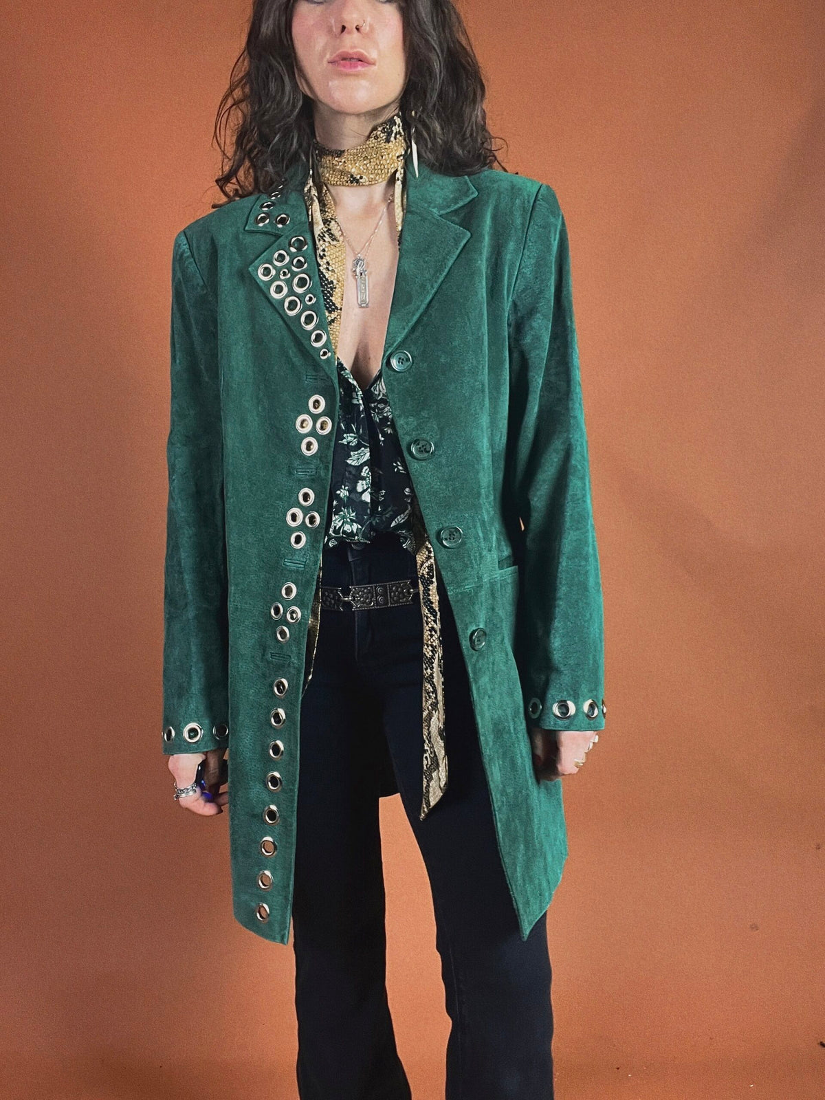 Woman posing in a green velvet coat with metallic button details, styled with a patterned scarf and black trousers.