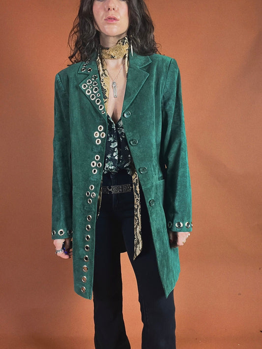Woman posing in a green velvet coat with metallic button details, styled with a patterned scarf and black trousers.