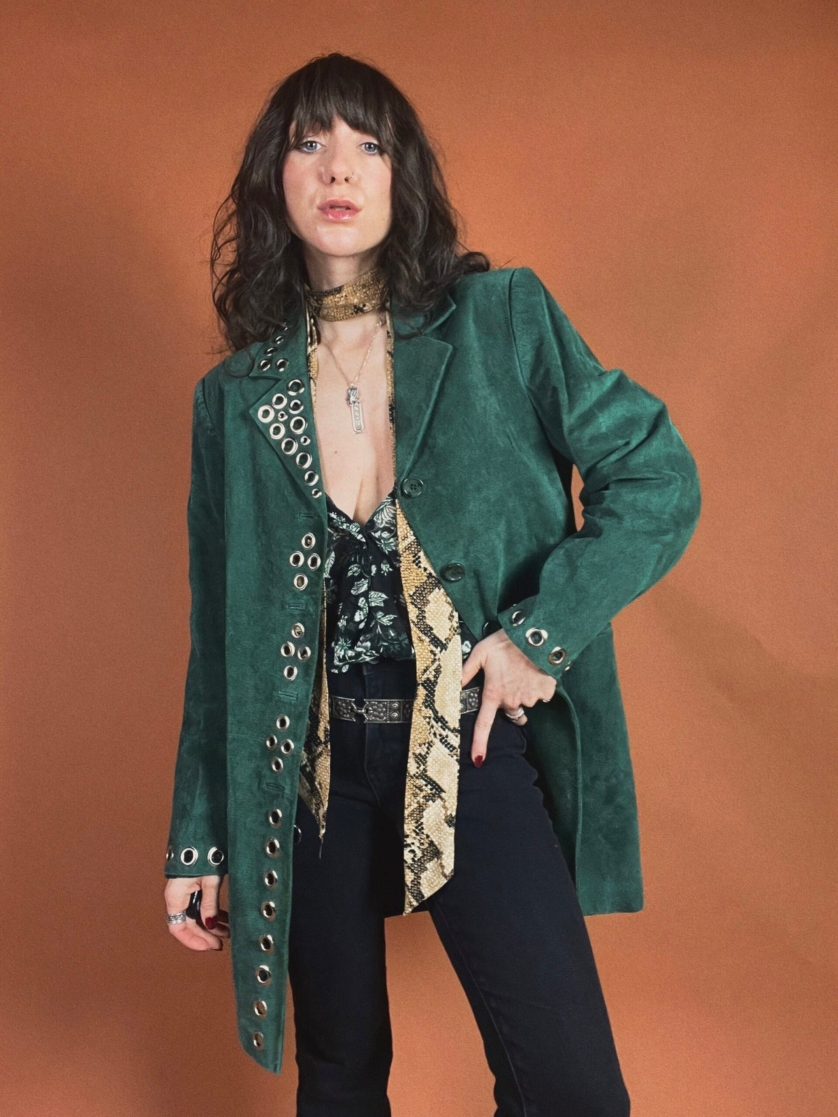 Woman posing in a stylish green jacket and black pants with a patterned blouse and accessories.