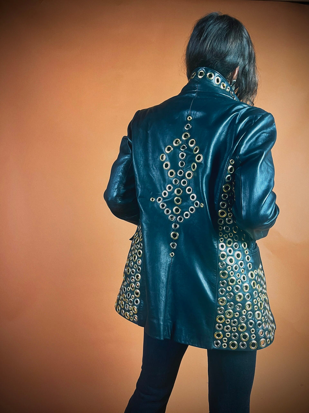 Person standing with their back to the camera, wearing a black leather jacket adorned with circular metal studs, against an orange background.