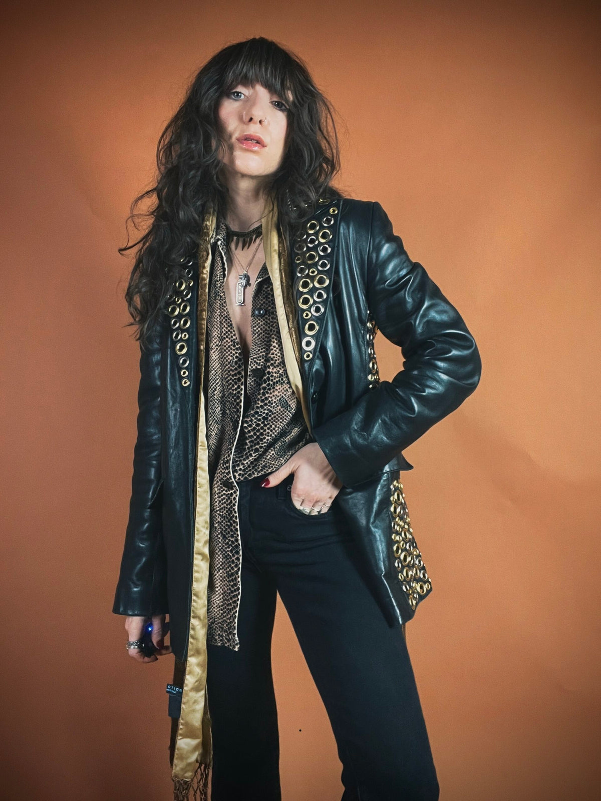 A person with long wavy hair wearing a black leather jacket, accessorized with multiple necklaces, against an orange backdrop.