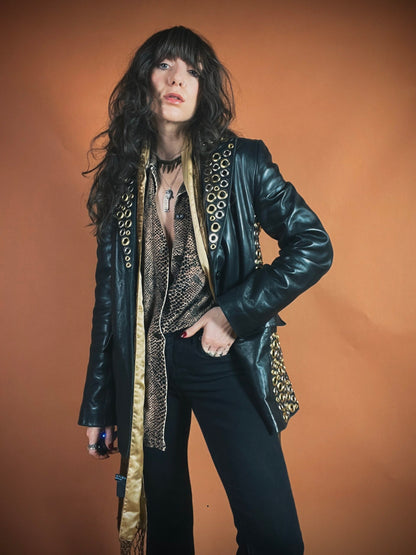 A person with long wavy hair wearing a black leather jacket, accessorized with multiple necklaces, against an orange backdrop.