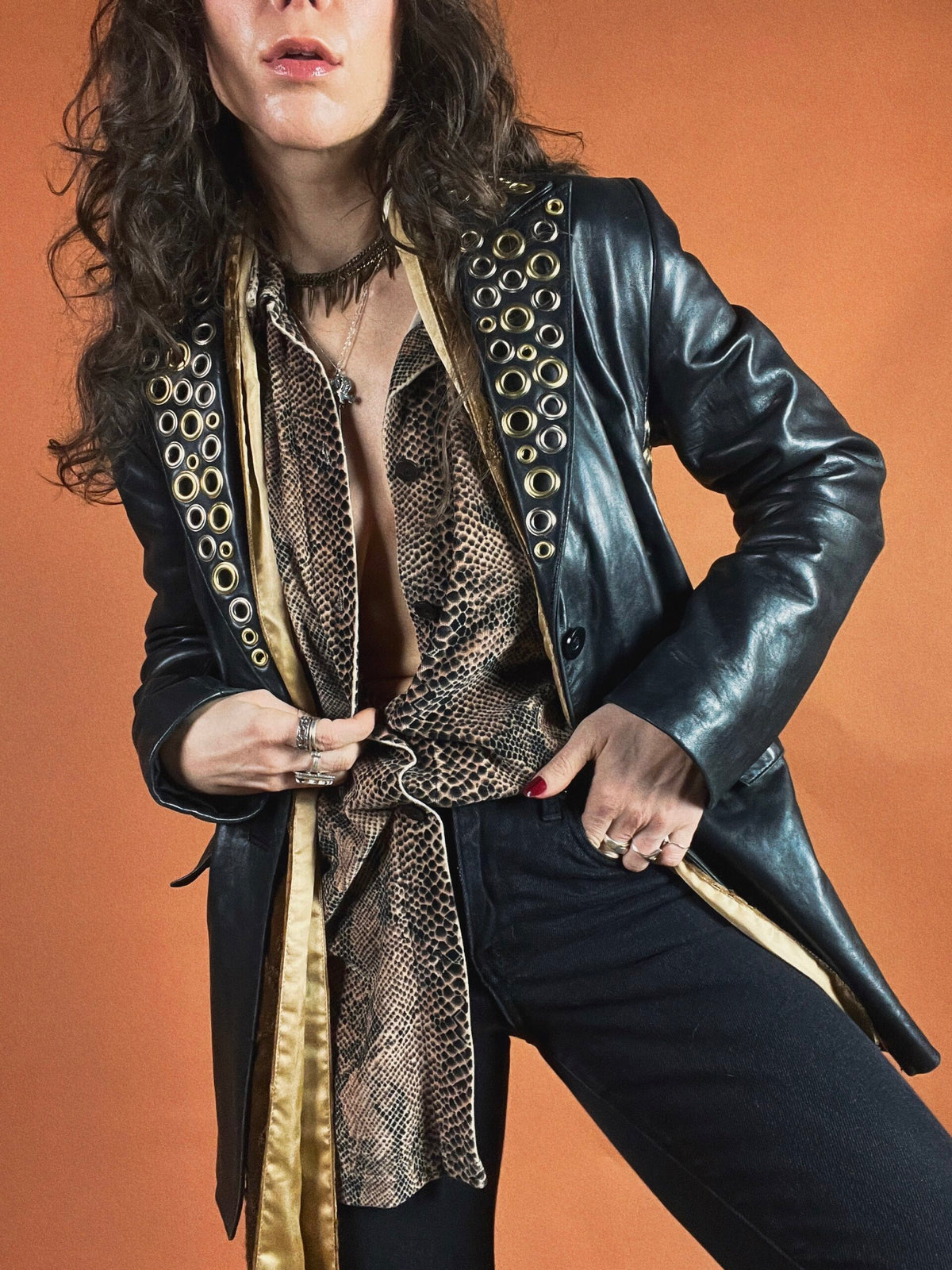 Woman in a black leather jacket with grommet detailing and snake-print blouse posing with hand on hip.