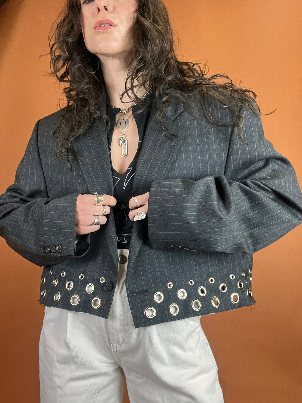 A person posing with hands on jacket lapels, wearing a pinstripe blazer and white trousers against an orange backdrop.