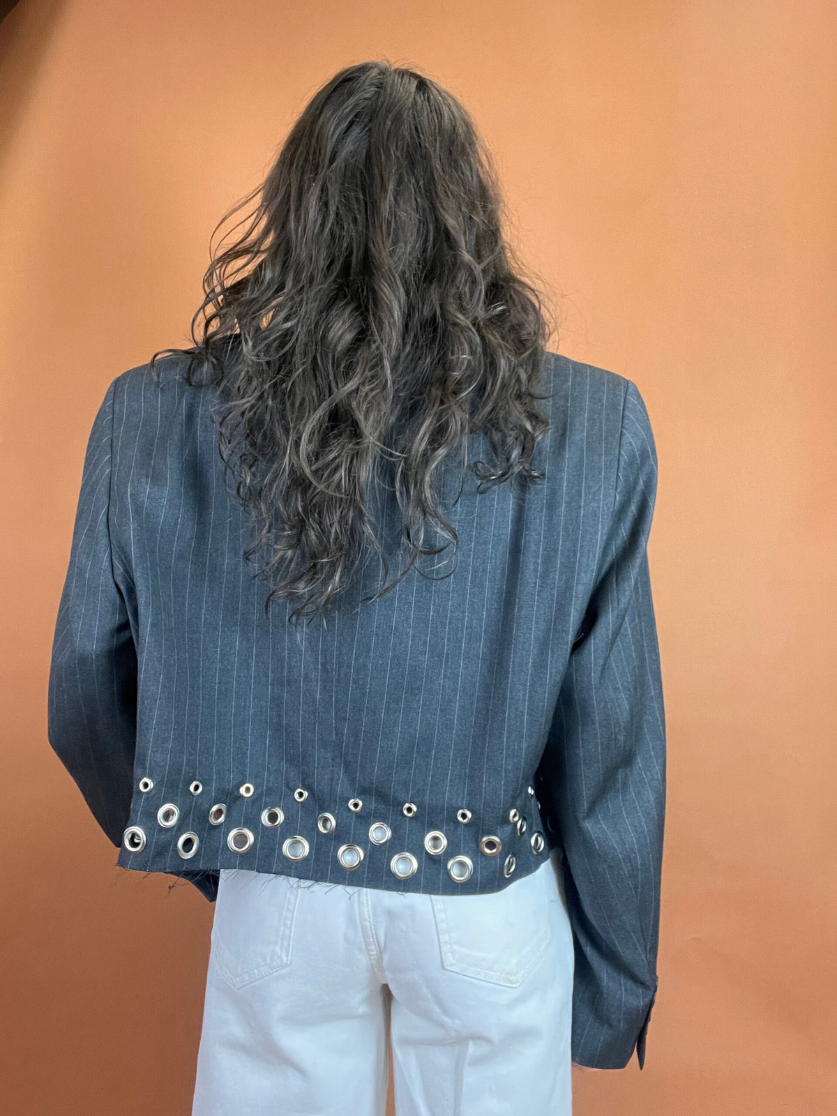 A person seen from behind wearing a pinstripe blazer with metal eyelet details on the hem and white pants against a tan background.