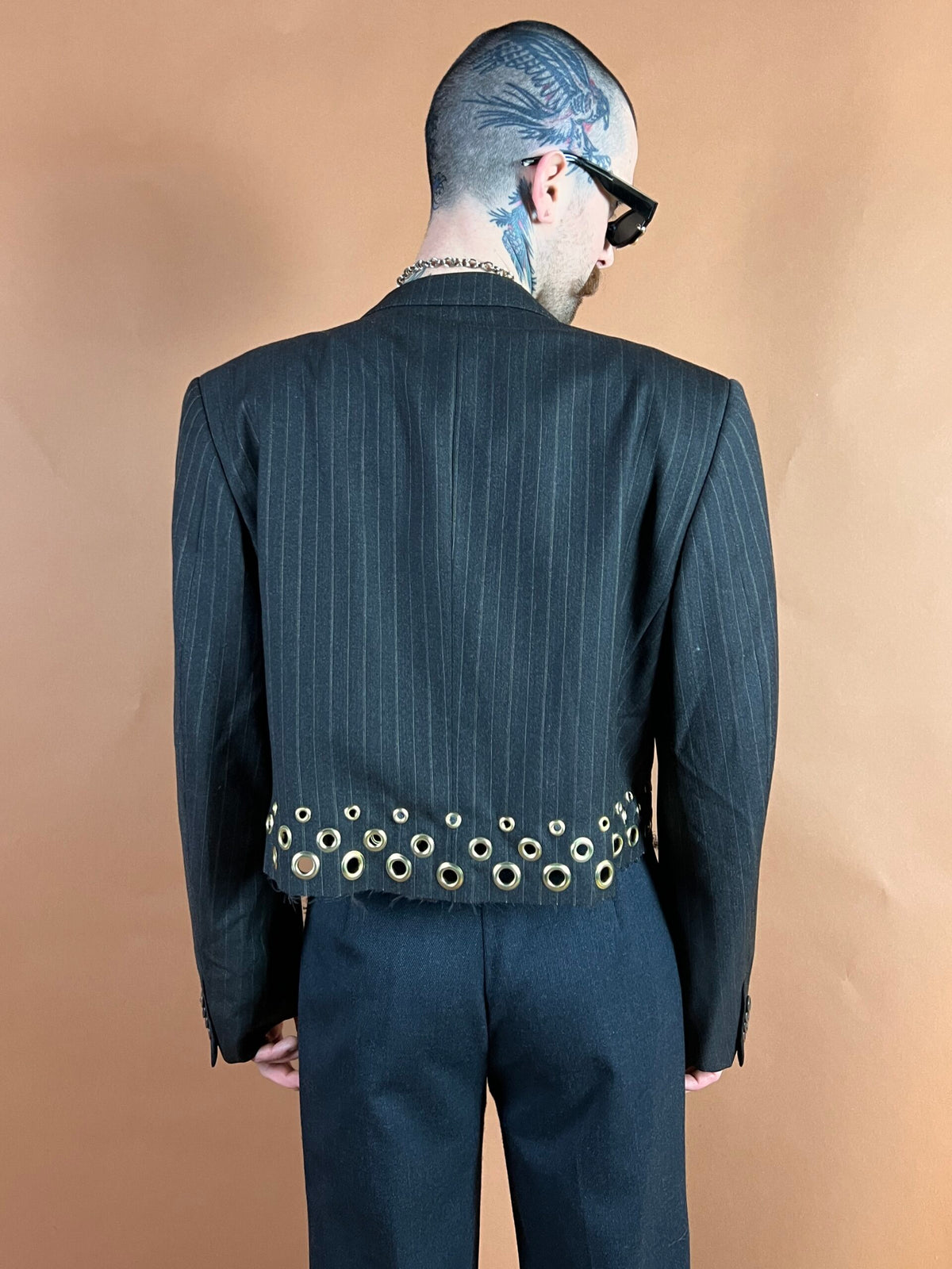 Man in a pinstripe suit with a tattoo on his head, wearing sunglasses and a studded belt, viewed from behind.