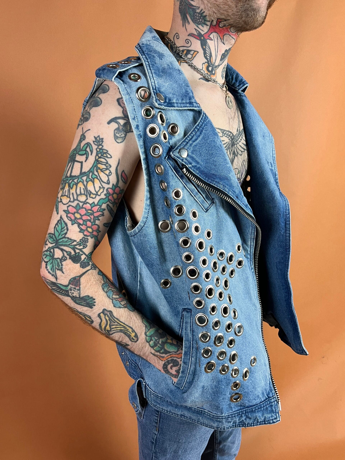 Person with tattoos wearing a studded denim vest against a brown background.