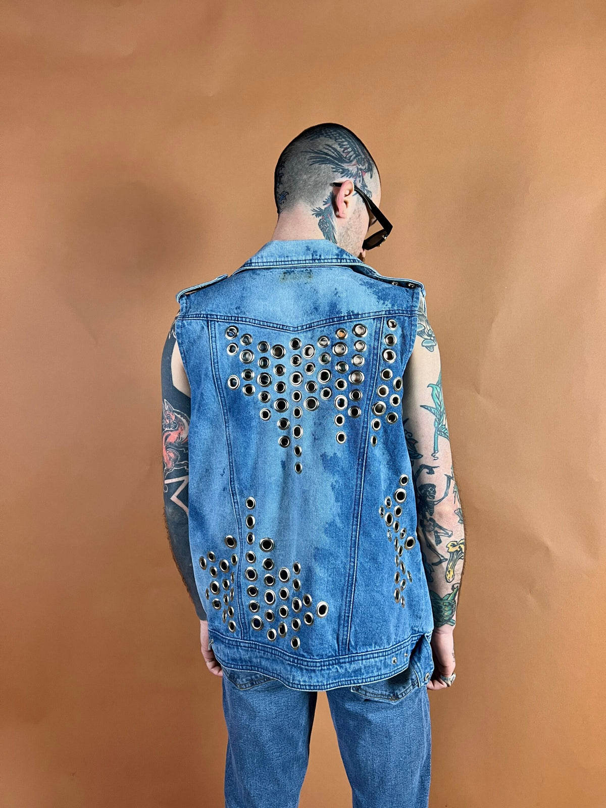 Man with tattoos wearing a customized denim vest with metal eyelets on a brown background.