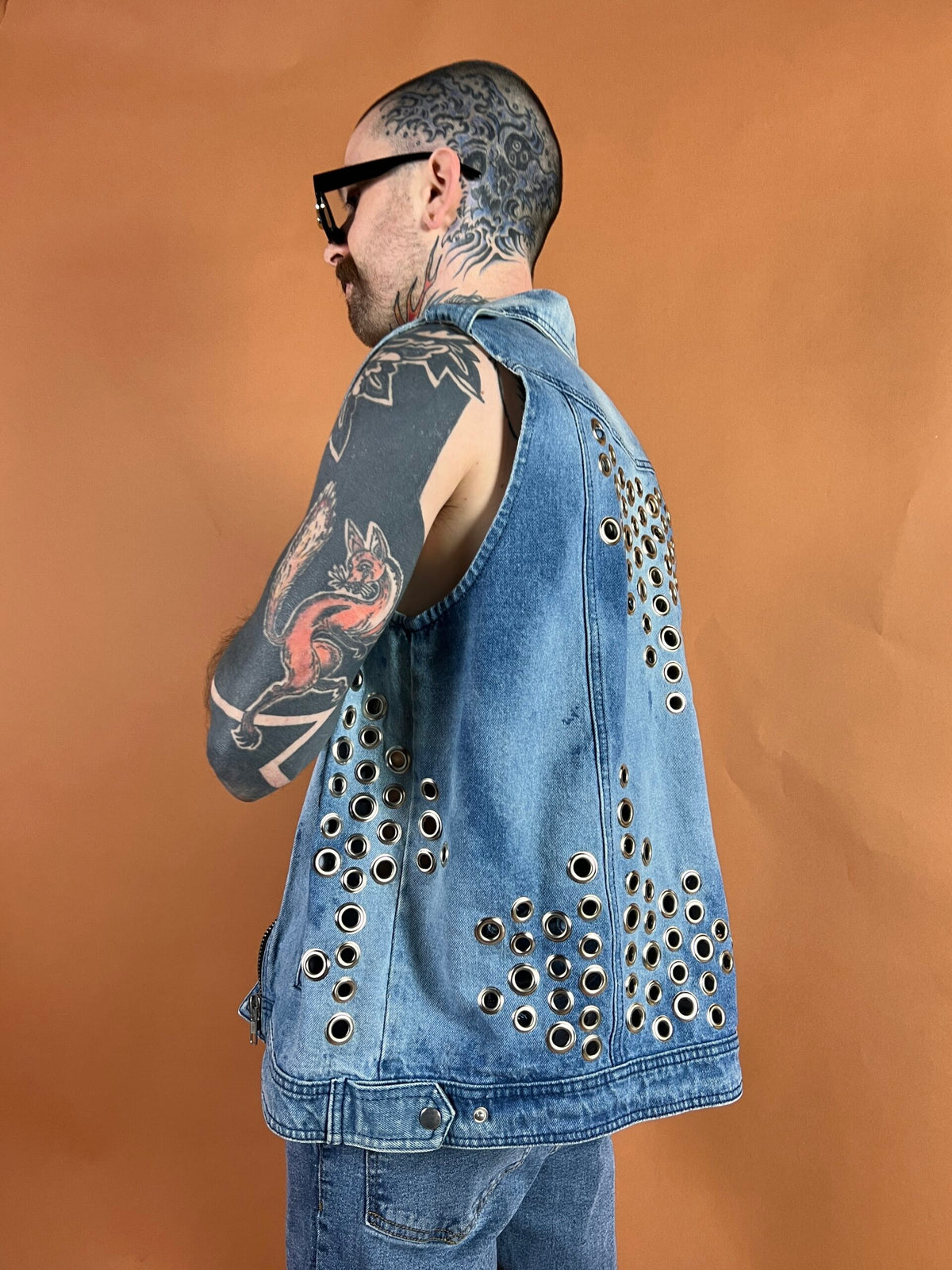 A person with a heavily tattooed neck and arms wearing a studded denim jacket against an orange background.