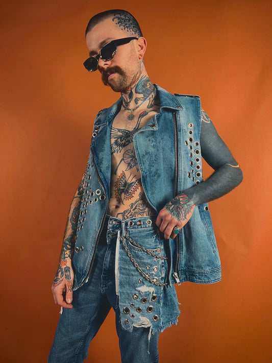 Man with tattoos wearing a sleeveless denim jacket and sunglasses against an orange background.