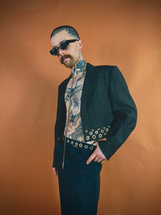 Stylish man in pinstripe suit and floral shirt posing confidently with sunglasses against an orange background.