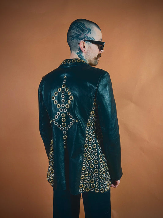A person with a tattooed head wearing a black leather jacket adorned with eyelets stands against a brown background.