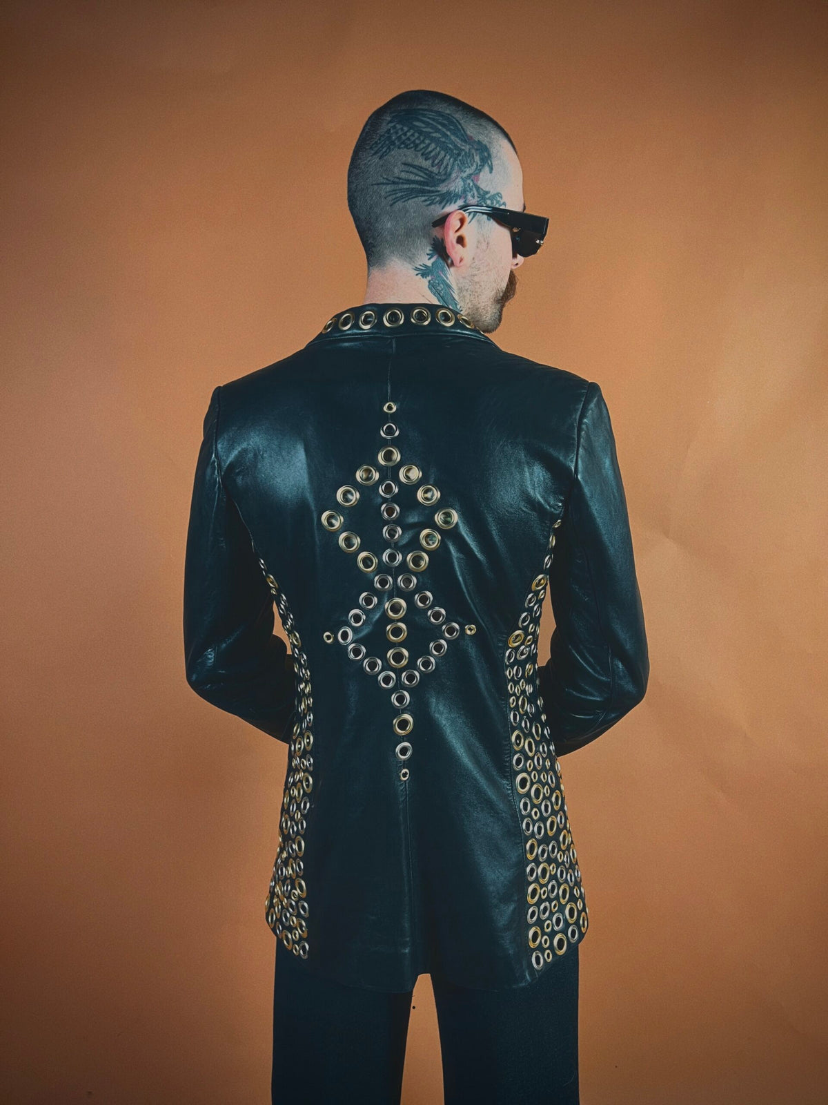 Man with a patterned undercut hairstyle wearing a leather jacket embellished with chains, facing away from the camera.