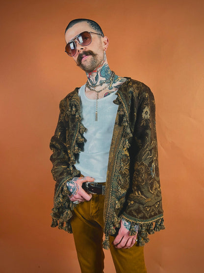 Man with tattoos and sunglasses wearing a patterned jacket and holding sunglasses.