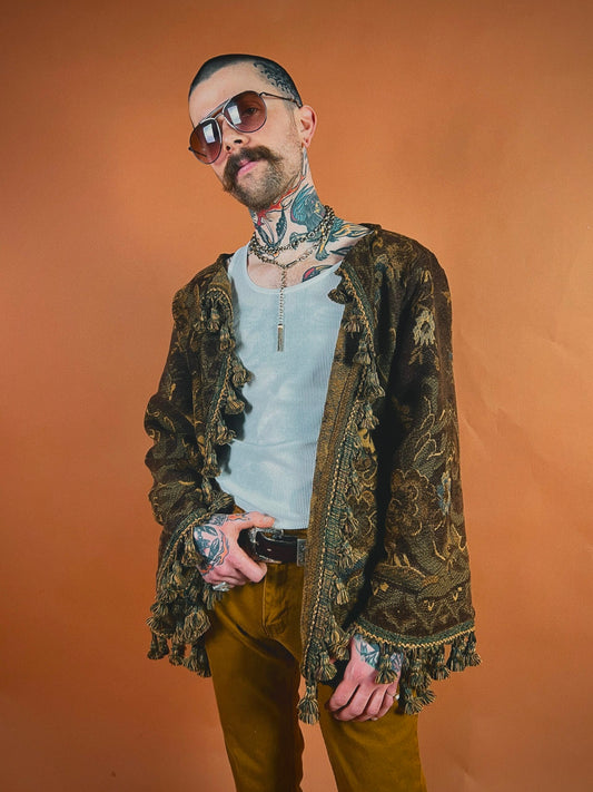 Man with tattoos and sunglasses wearing a patterned jacket and holding sunglasses.