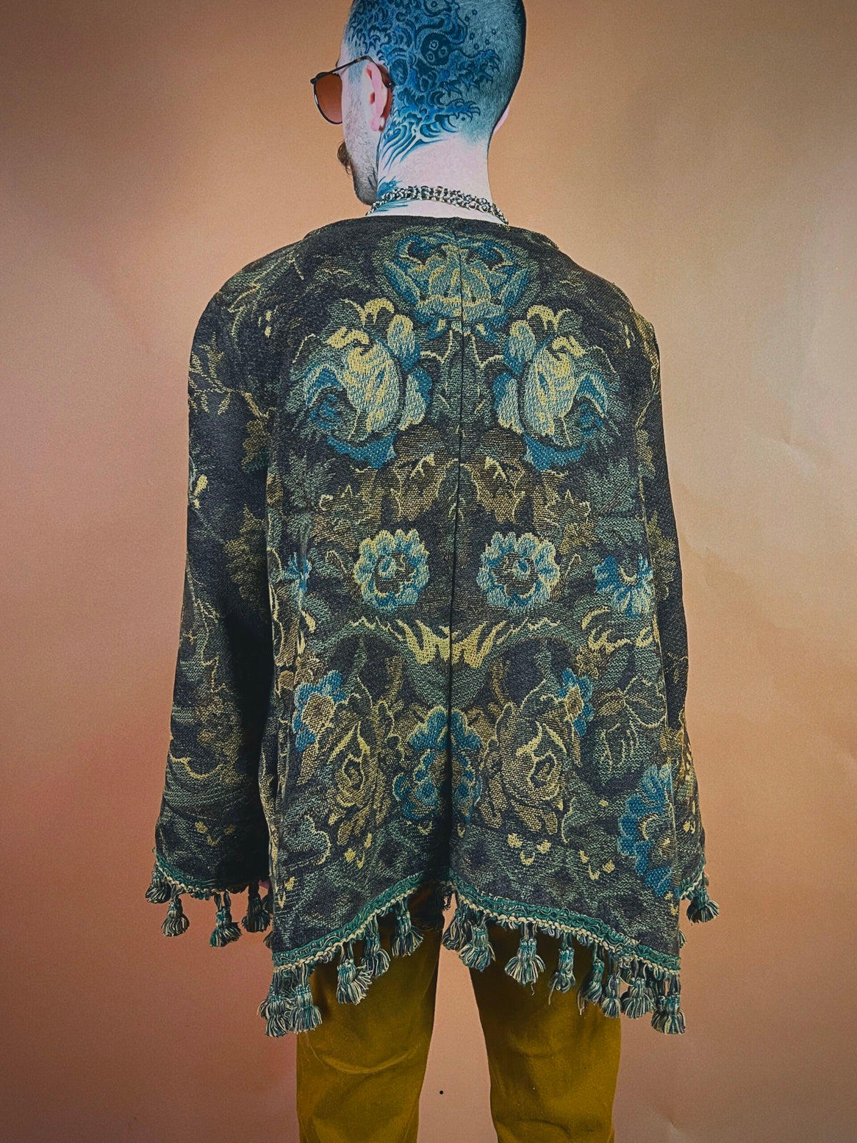 A person standing with their back to the camera wearing a floral patterned jacket with fringe details and mustard-colored trousers. visible tattoos are present on the person's head and neck.