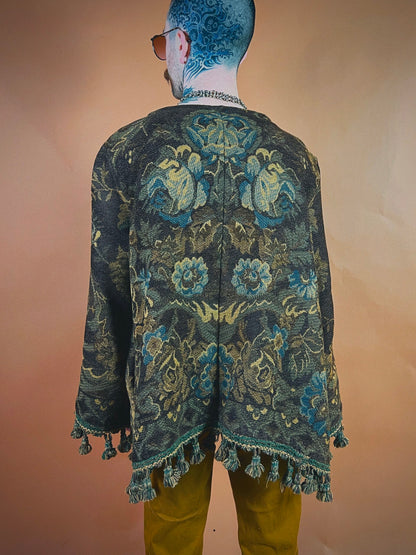 A person standing with their back to the camera wearing a floral patterned jacket with fringe details and mustard-colored trousers. visible tattoos are present on the person's head and neck.