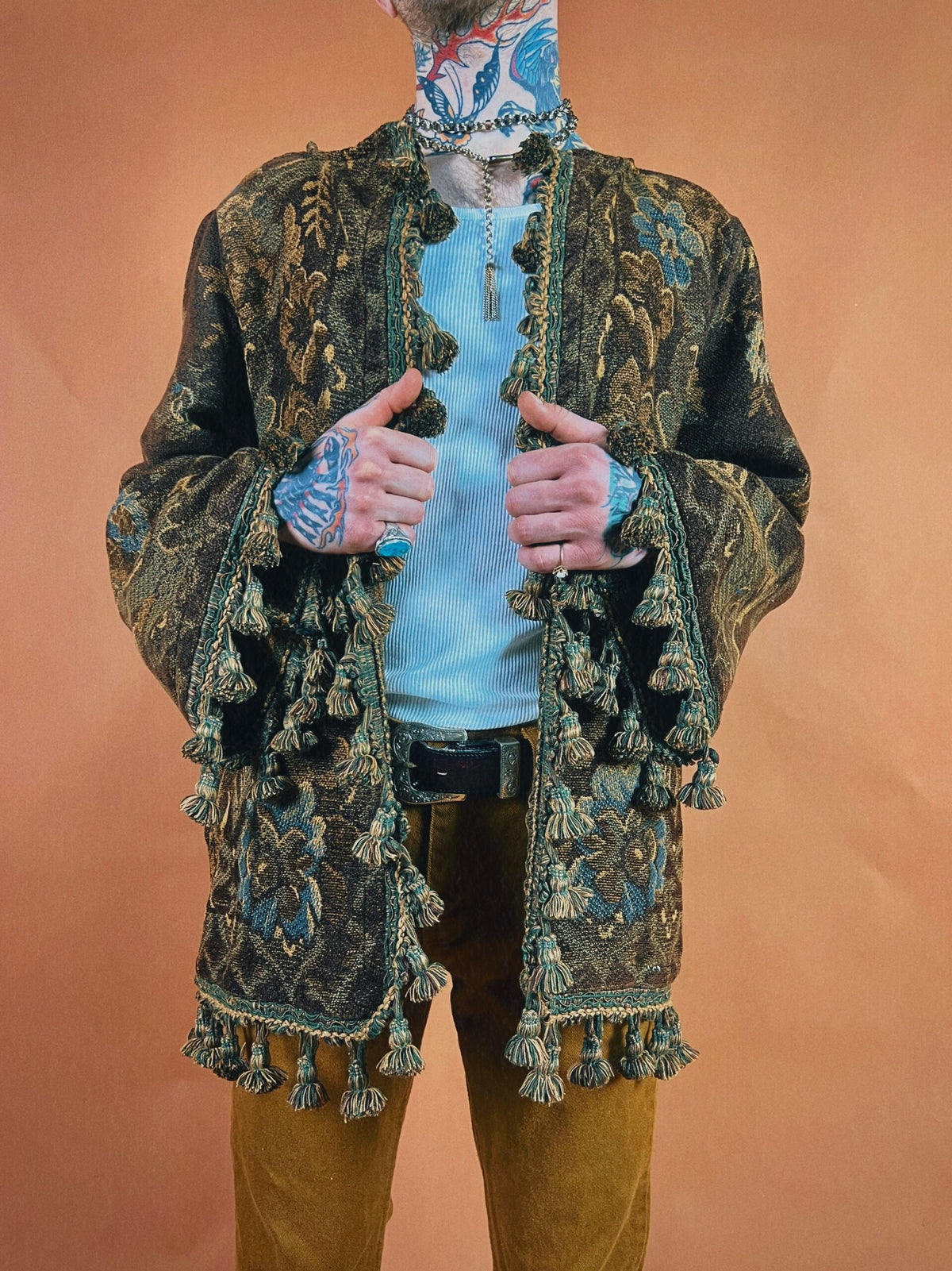 A person with tattoos wearing an ornate jacket with tassel details stands against an orange backdrop, face obscured from view.