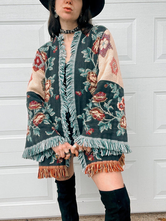 A person in a floral patterned poncho and black hat standing in front of a white door.