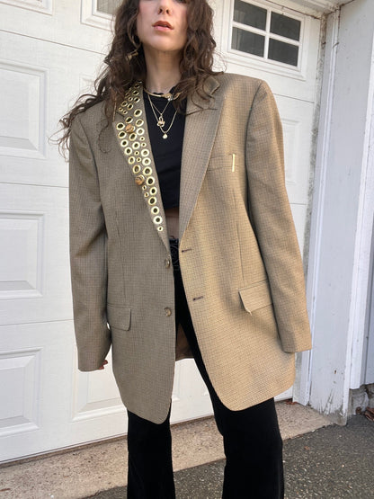 A person wearing a herringbone blazer over a black top, adorned with a long golden necklace, paired with black trousers, standing in front of a white garage door.