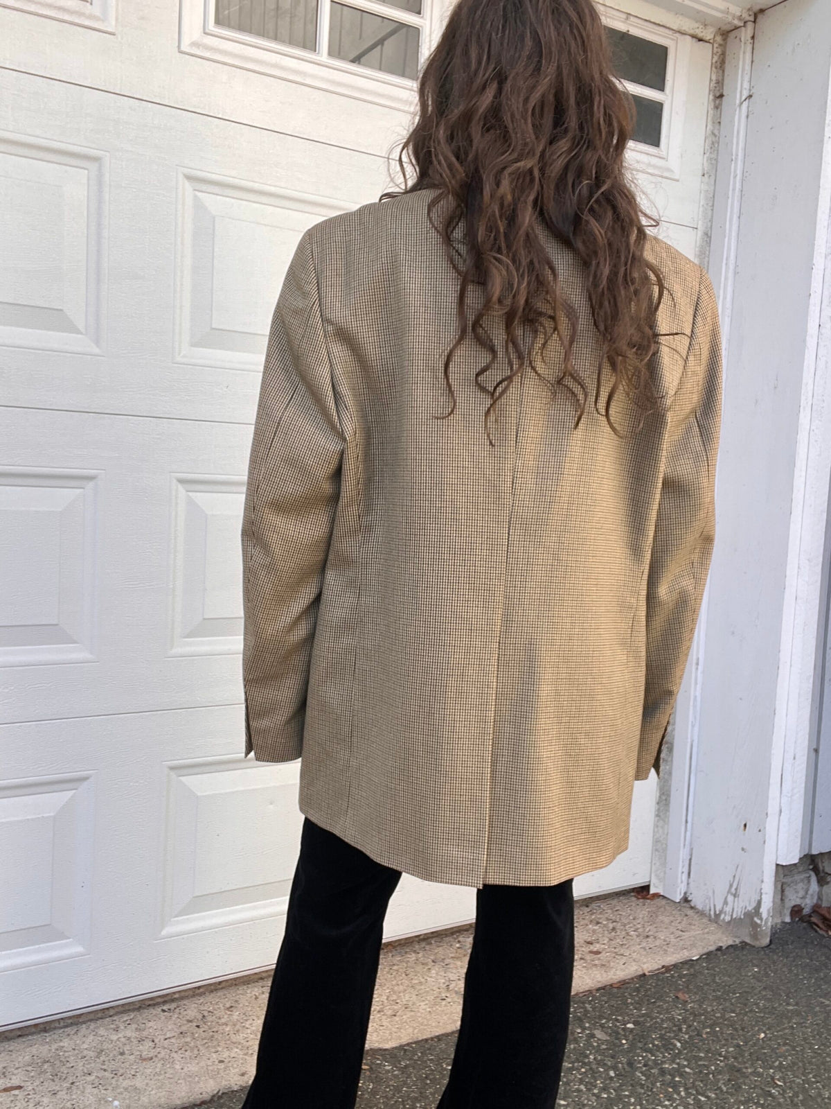 Person standing with their back to the camera wearing a beige blazer and black pants.