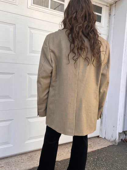 Person standing with their back to the camera wearing a beige blazer and black pants.