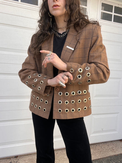 A person wearing a plaid blazer with eyelet details and black pants, accessorized with a chunky chain necklace and multiple rings.